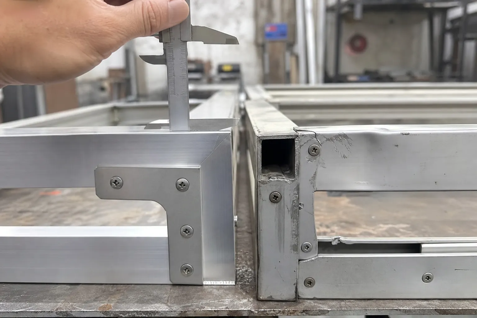 Cách Tối Ưu Chi Phí Tủ Bếp Nhôm Mà Không Giảm Chất Lượng Quan Trọng 2 Ảnh chụp cận cảnh hai mẫu khung tủ bếp nhôm đặt cạnh nhau trên bàn xưởng bên trái là khung nhôm dày, vuông góc chuẩn, liên kết bằng ốc vít và ke góc chắc chắn, mối hàn gọn; bên phải là khung nhôm hộp mỏng, mép bị móp nhẹ, lỗ vít lệch, mối nối hở và xước sơn; có tay người thợ cầm thước kẹp đo độ dày thanh nhôm để so sánh trực quan