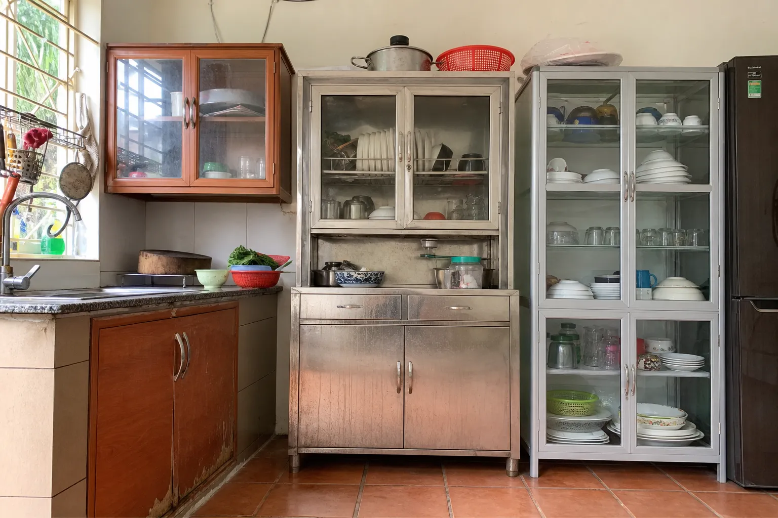 Chạn Bát Và Tủ Bát Đĩa Nhôm Kính: Hướng Dẫn Toàn Diện 1 Chạn Bát Và Tủ Bát Đĩa Nhôm Kính Không gian bếp gia đình Việt Nam với ba mẫu tủ đặt cạnh nhau để so sánh thực tế một tủ gỗ công nghiệp có mép cánh hơi trương gần chậu rửa, một tủ inox có vài vệt nước và điểm ố nhẹ ở chân, một tủ nhôm kính sạch sẽ với khung nhôm màu xám mờ và cánh kính trong, ánh sáng tự nhiên chiếu ngang làm lộ rõ bề mặt vật liệu, góc chụp tầm mắt, cảm giác đời thường chứ không dàn dựng quảng cáo